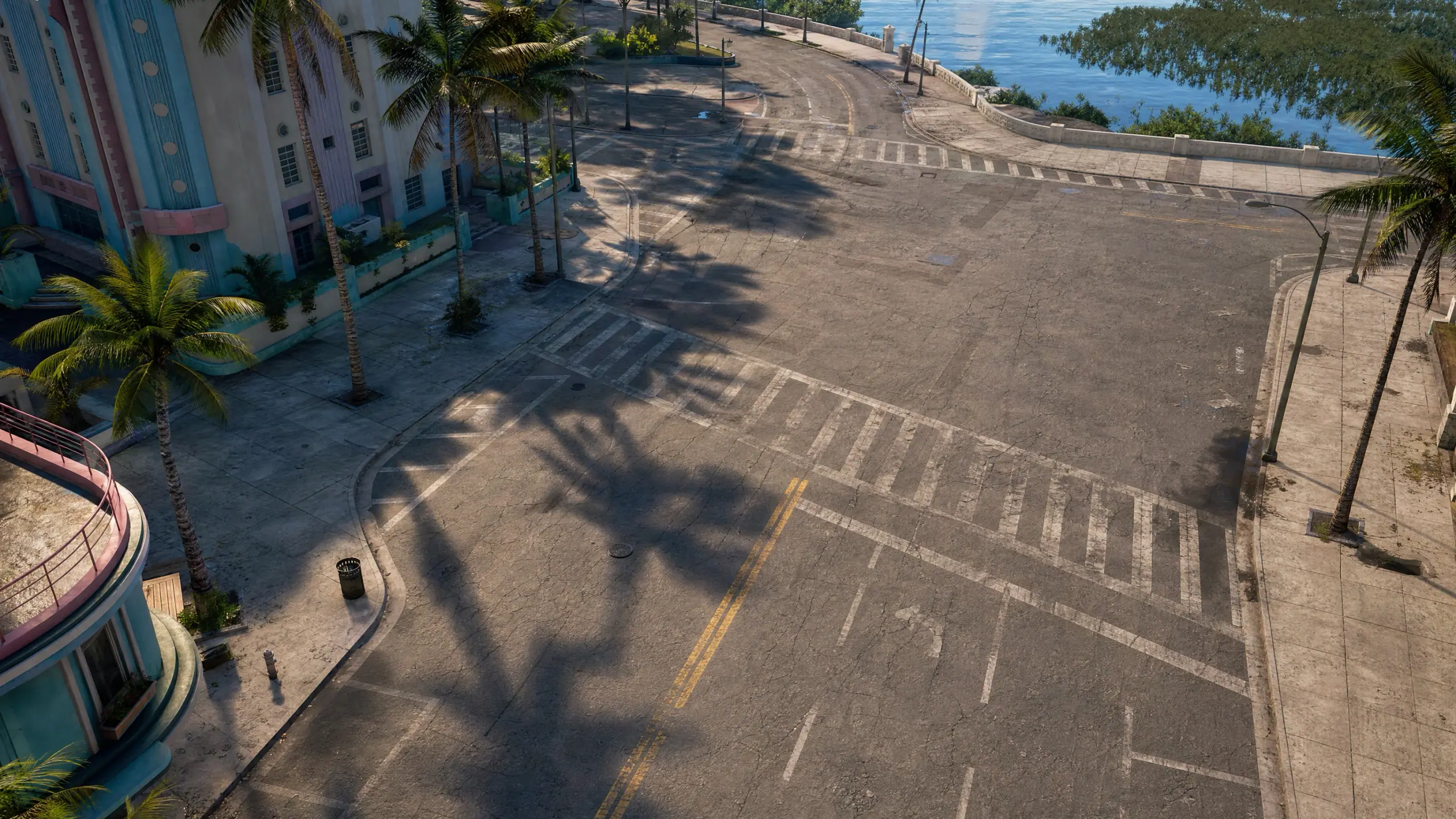 Our own overhead render of a quiet road junction: worn asphalt and faded lane markings across most of the frame, a curved pastel building and palms throwing long shadows at one edge, and water beyond the seawall.
