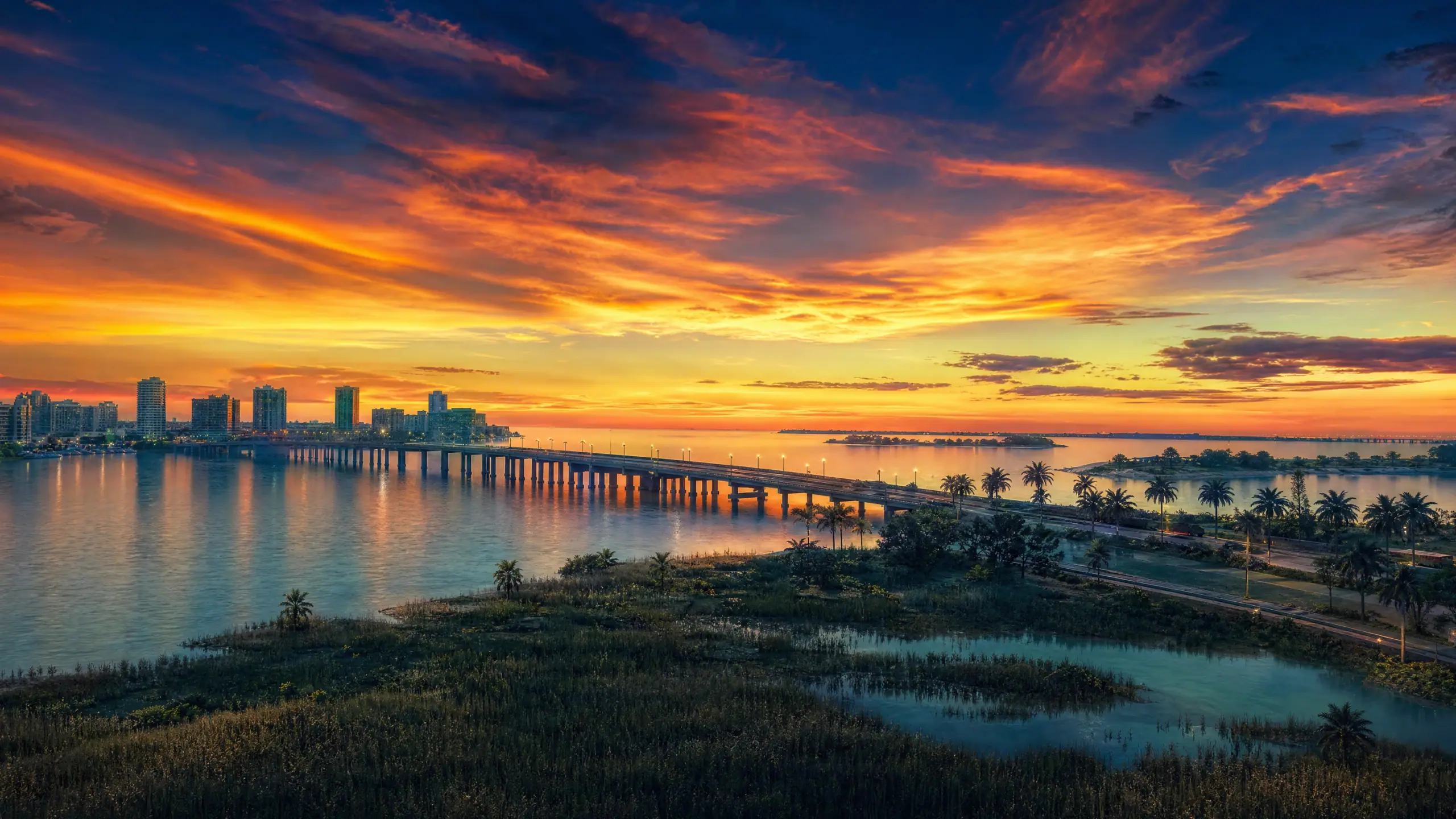 Our own dusk render over a wide bay: banded orange and deep blue cloud above a low city skyline, a long causeway crossing the water beneath it and dark mangrove scrub across the foreground.